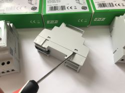 Śrubokręt rozchyla plastikową obudowę białego modułu elektrycznego na tle zielonych opakowań.