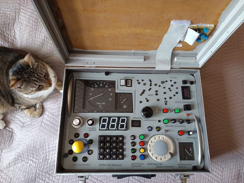 Voyager 3 Control Center – DIY Control Panel in a Suitcase A suitcase with a homemade electronic control panel inspired by a space station toy, standing on a bed, with a cat lying next to it.