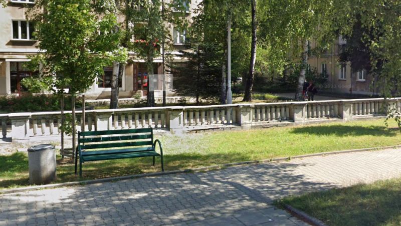 Parkowy skwer z zieloną ławką, trawnikiem, drzewami i niską balustradą przy budynkach