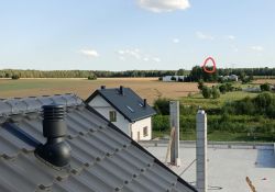 Widok z dachu domu na wiejską okolicę z polami i lasem. W dali widoczny las z czerwoną obwódką.