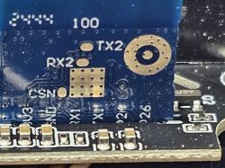 Close-up of a blue PCB with labels TX2, RX2, GND and gold solder pads visible.