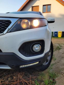 Przód białego samochodu KIA Sorento II z włączonymi światłami.