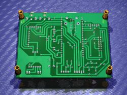 Bottom view of a green PCB with many traces and through-holes, plus four gold mounting standoffs