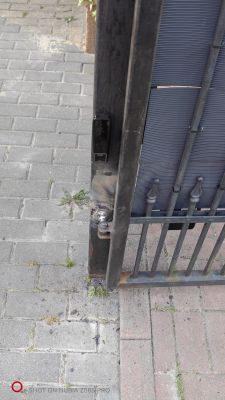 Zbliżenie na dolną część metalowej bramy przesuwnej z zamontowaną fotokomórką oraz widocznym elementem zamka na tle chodnika z kostki brukowej.