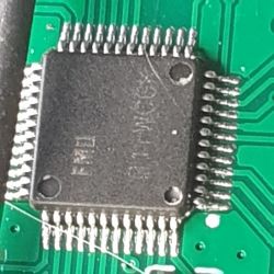 Close-up of an integrated circuit on a green circuit board with visible pins on the sides.