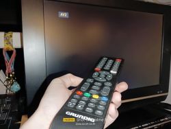 Dłoń trzyma uniwersalny pilot Grundig przed telewizorem z napisem „AV2” na ekranie