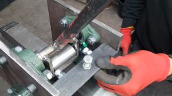 Close-up of a metal mechanism with bearing housings; red-gloved hand points at a bolt