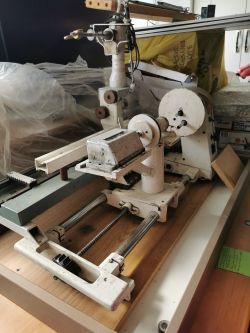Stara maszyna mechaniczna na metalowej ramie z elementami przesuwającymi się na prowadnicach, umieszczona na drewnianej podstawie.