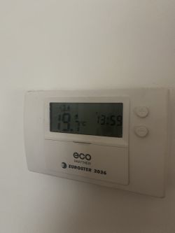 Cyfrowy termostat na ścianie z wyświetlaną temperaturą 19,7°C i godziną 13:59.