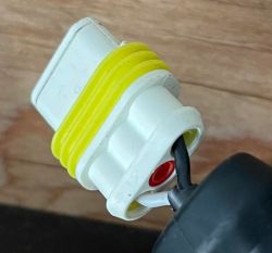 Zbliżenie na biały złącze elektryczne z żółtą uszczelką, na tle drewnopodobnym.