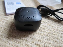 Miniature Black Speaker on Carpet A small, black, square speaker or electronic device with vent holes and a slot, placed on a beige carpet.