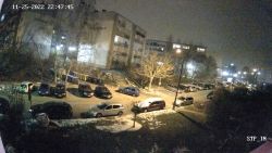 Widok z kamery nocą na osiedle z blokami mieszkalnymi, zaparkowanymi samochodami i oświetleniem ulicznym.