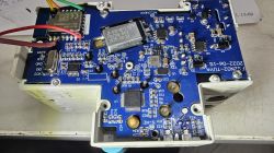 Exposed blue PCB with SMD components, wires, and a shielded module inside a white enclosure