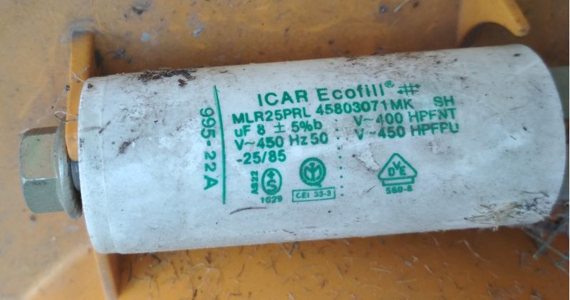 Zbliżenie na biały kondensator rozruchowy marki ICAR Ecofill z oznaczeniami technicznymi, zamontowany w obudowie kosiarki.