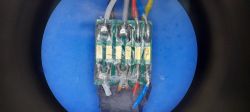 Zbliżenie płytki PCB z przylutowanymi przewodami na niebieskim tle, w okrągłym kadrze.