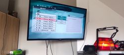 Telewizor na ścianie z menu „MUSIC” i listą utworów; pod spodem zwisają kable