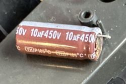 Uszkodzony kondensator elektrolityczny 10µF 450V leżący na metalowej powierzchni, widoczne oznaczenia parametrów.