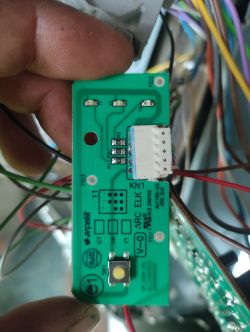 Zbliżenie na zielony moduł elektroniczny z przyciskiem, przewodami i złączem, trzymany w dłoni.