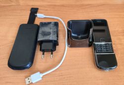Telefon Nokia z klawiaturą numeryczną oraz stacja dokująca, ładowarka USB i czarne etui na drewnianym blacie