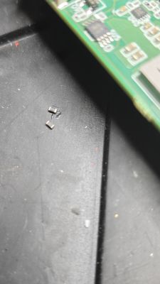 Dwa małe elementy SMD na czarnej powierzchni obok fragmentu zielonej płytki PCB