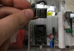 Electronic module held in hand against a breadboard with components in the background.