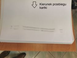Arkusz papieru z szarymi smugami tonera w dolnej części i napisem „Kierunek przebiegu kartki” ze strzałką
