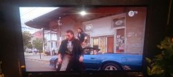 Scena z filmu na ekranie telewizora z logiem TVN HD, pokazująca dwóch mężczyzn przy niebieskim kabriolecie na tle budynku.