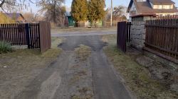 Otwarta brama skrzydłowa na posesję z widokiem na drogę i domy w tle.