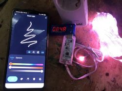 Smartphone with LED light control app, meter displaying 0.84A, and lit LED string lights.