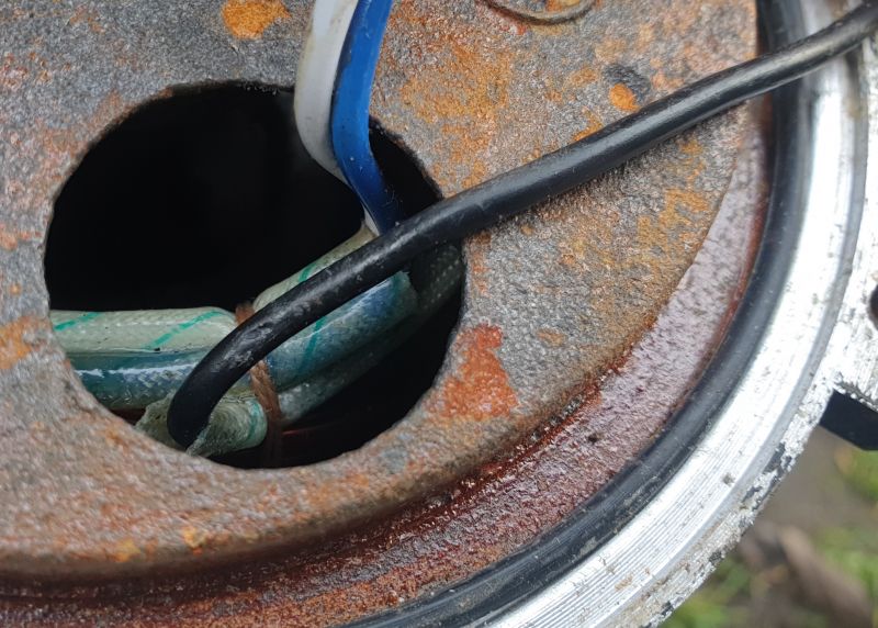 Zbliżenie na kabel czarny i inne przewody przechodzące przez otwór w zardzewiałej metalowej powierzchni.