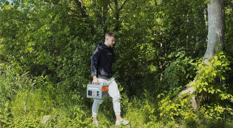 Młody mężczyzna idzie przez zielony las, trzymając w ręku przenośną stację zasilania.