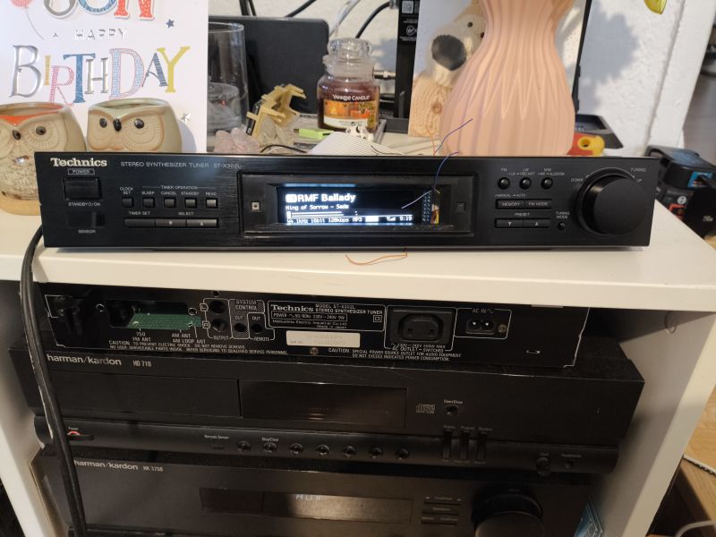Technics ST-X302L tuner with lit display, on a shelf above Harman/Kardon audio components