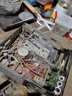 Wnętrze starego radia z widoczną płytką drukowaną, przewodami i mechanizmem kasety, na zakurzonej powierzchni roboczej.