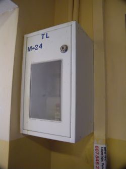 Biała metalowa skrzynka z okienkiem i zamkiem, oznaczona literami TL i M-24, zamocowana na ścianie klatki schodowej.