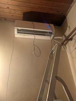 Wall-mounted air conditioner showing “23”, with hanging wires and a ladder leaning against the wall.