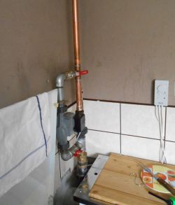 Zbliżenie na część instalacji hydraulicznej z rurami miedzianymi i zaworami, obok drewniana deska na blacie i termostat.