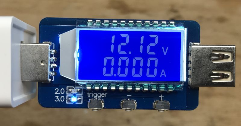 QC tester with blue LCD display showing 12.12 V and 0.000 A, connected to a charger via USB port.