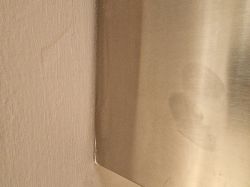 Zbliżenie na luźno dopasowaną metalową osłonę przylegającą do ściany, z widoczną szczeliną w rogu.