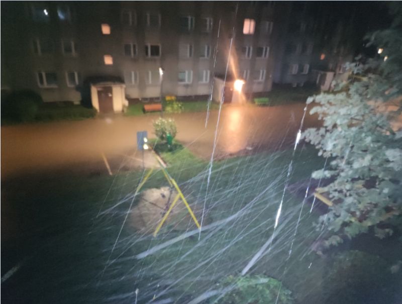 Zdjęcie pokazujące zalany teren przed blokiem mieszkalnym podczas deszczu.