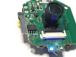 Close-up of a camera circuit board with lens, T25S80 flash chip, and a lit blue LED.