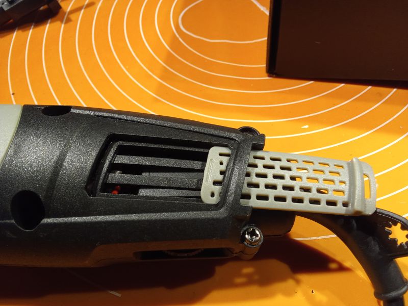 Zbliżenie na czarną szlifierkę z wysuniętą kratką wlotu powietrza, umieszczoną na pomarańczowym tle.