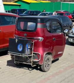 Bordowy trzykołowy mikroauto z tyłu z zamocowanym wózkiem i dużym urządzeniem na stelażu, na parkingu.