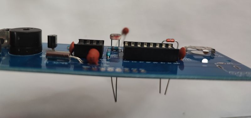 Side view of a blue PCB with a buzzer, DIP sockets, capacitors, and a coin-cell battery holder