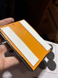 A hand holds an orange rectangular piece with two white adhesive strips on top