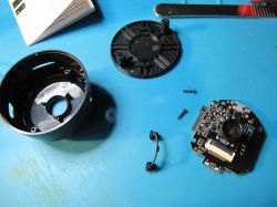 Disassembled Camera with Components Disassembled camera with electronic circuit board, lens, and casing parts on a blue surface.