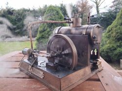 Stara maszyna parowa z wirującym kołem, ustawiona na drewnianym stole w ogrodzie.