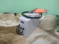 Małe urządzenie elektroniczne w białej obudowie z wyświetlaczem LED i dwoma pokrętłami, z przymocowanymi przewodami oraz uchwytem z pomarańczową osłoną.