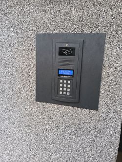 Domofon z klawiaturą numeryczną zamontowany na ścianie pokrytej szarym tynkiem.