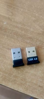 Dwa adaptery Bluetooth USB, jeden opisany jako CSR 4.0, leżą na drewnianym blacie.