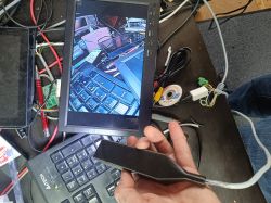 Osoba trzymająca czujnik lub kamerę przed małym monitorem na biurku z różnymi elektronicznymi przedmiotami, w tym klawiaturą i kablami.